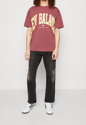 Maroon t-shirt med fed gul "NEW BALANCE" tekst, parret med sorte jeans og hvide sneakers med sorte striber. Casual pasform og design.