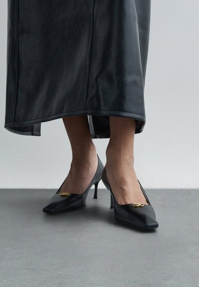 Kobieta nosząca czarne czółenka na niskim obcasie z złotymi ozdobami, zestawione z czarną skórzaną spódnicą midi, stojąca na szarej podłodze.