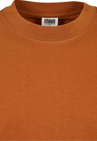 T-shirt col rond couleur rouille avec étiquette visible "Urban Classics Organic" à l'intérieur du col, montrant la texture du tissu et les détails des coutures.