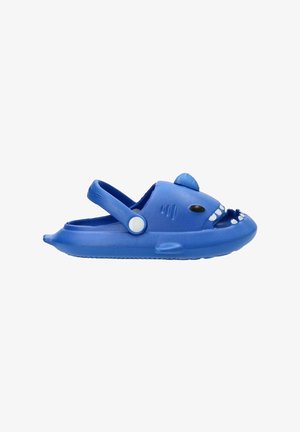 Zuecos de plástico azules con forma de tiburón, boca sonriente, acentos blancos, correa lateral y forma redondeada. Suela texturizada para mayor agarre.