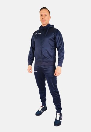 Marine trainingspak van soepel materiaal met een rits hoodie en elastische boorden. Voorzien van een wit logo op de borst en sportschoenen met contrasterende veters.