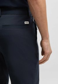 Navy blue trousers made from smooth fabric, featuring a back pocket with a "BOSS" label and a side zipper detail. Hand positioned near the pocket.