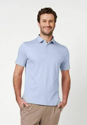 Homme souriant portant un polo bleu clair à manches courtes et un pantalon beige, les mains dans les poches, sur fond blanc.