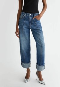 Jeans de mezclilla azul claro con una cintura alta, corte relajado, puños enrollados y detalles de costura en contraste, combinados con tacones metálicos.