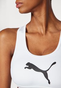 Hvit sports-BH med en svart Puma-logo på brystet. Har en rund utrigning og glatt, elastisk materiale.
