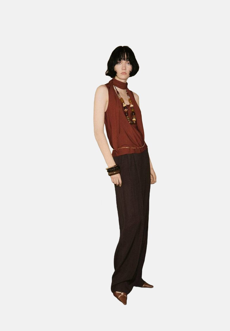 Top senza maniche di colore ruggine con design drappeggiato, abbinato a pantaloni larghi scuri. Accessori includono una collana multicolore di perline e braccialetti sovrapposti.