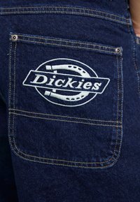 Denim-kangas, tummansininen väri, jossa on takatasku, jossa on valkoisella brodeerattu "Dickies"-logo ja kontrastivärinen keltainen tikkaus.