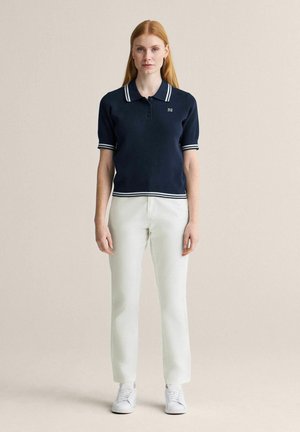 Kvinde iført en marineblå kortærmet strikket polo med hvide striber, hvide bukser og hvide sneakers stående foran en ensfarvet baggrund.