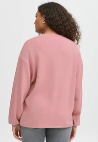 Felpa rosa chiaro con spalle basse, maniche lunghe e una consistenza soffice, abbinata a pantaloni grigi con una texture particolare. Nessun motivo o hardware visibile.