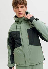 Green and black zip-up jacket with a hood, featuring large pockets, adjustable drawstrings, and contrasting panels. Smooth texture.