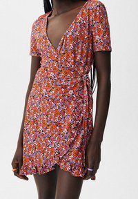 Rotes Blumenkleid mit kurzen Ärmeln, tiefem V-Ausschnitt, gerafftem Seitendetail und einem rüschierten Saum. Der Stoff hat eine weiche Textur.