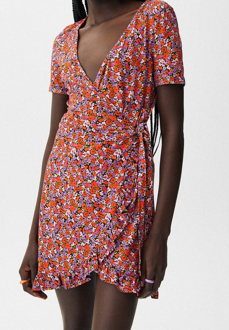 Rotes Blumenkleid mit kurzen Ärmeln, tiefem V-Ausschnitt, gerafftem Seitendetail und einem rüschierten Saum. Der Stoff hat eine weiche Textur.