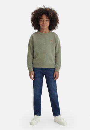 Vaikas su garbanotais plaukais, vilkintis alyvuogių žalios spalvos sportinį megztinį, mėlynas džinsas ir baltus sportinius batelius, stovintis veidu į priekį ant balto fono.