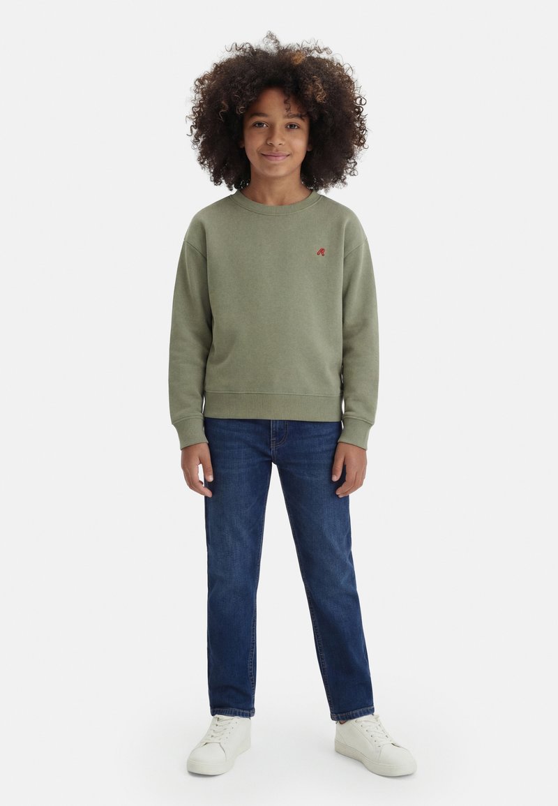 Dítě se zvlněnými vlasy, které má na sobě olivově zelenou mikinu, modré džíny a bílé tenisky, stojí čelem čelem na bílém pozadí.