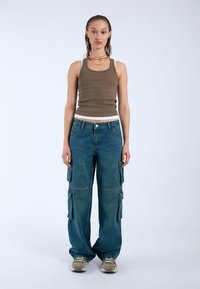 Top sin mangas de color marrón con líneas y pantalones cargo de mezclilla azules, holgados, con bolsillos con cremallera. Modelo lleva zapatillas neutras, de pie frente a un fondo liso.