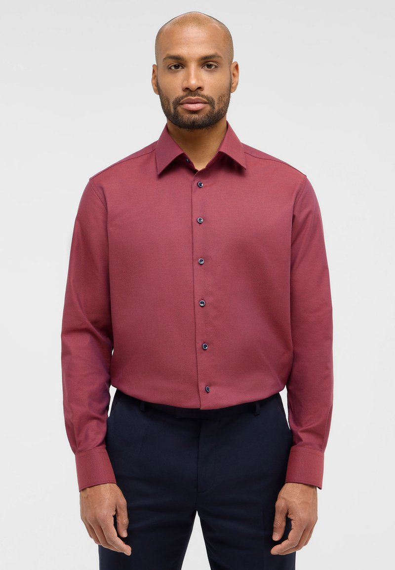 Una camicia a maniche lunghe color marrone scuro con un design testurizzato, chiusura frontale con bottoni, bottoni scuri a contrasto e colletto a punta, abbinata a pantaloni scuri.