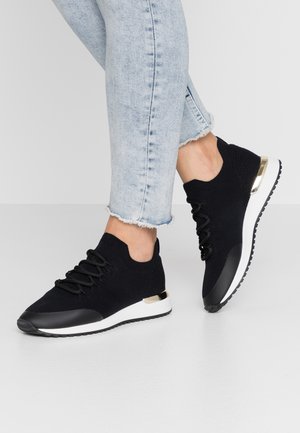 Osoba nosząca jasnoniebieskie dżinsy z postrzępionymi nogawkami oraz czarne sneakersy na sznurowadła z białą podeszwą i złotymi akcentami na pięcie, stojąca na szarej podłodze.
