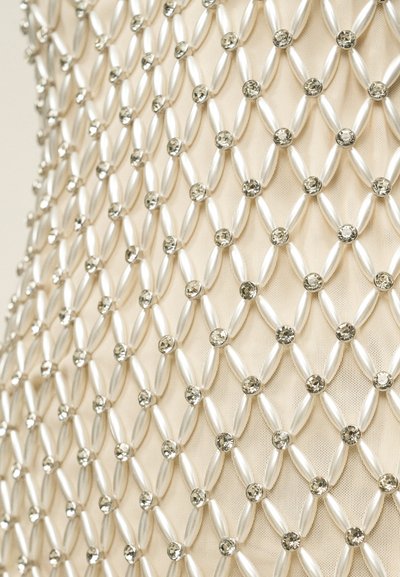 Μπεζ ύφασμα διακοσμημένο με πλέγμα από ασημένιες επιμηκυμένες χάντρες και μικρές στρογγυλές στρας που σχηματίζουν ρόμβους.