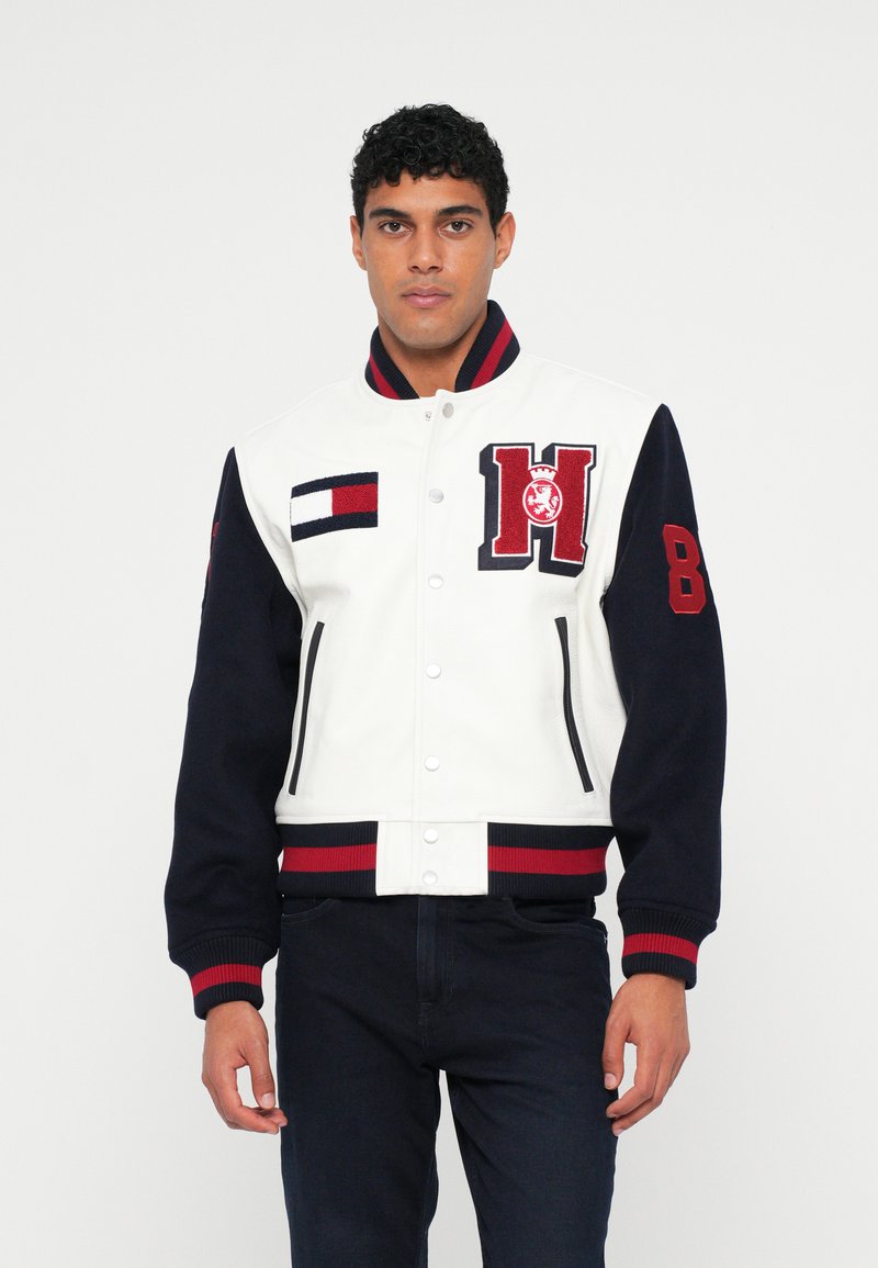 Giacca varsity in pelle bianca e lana blu navy con dettagli rossi e blu navy, dotata di un logo ricamato e polsini a righe. Chiusura frontale con bottoni a pressione.