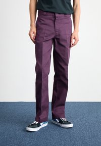 Pantaloni din bumbac mov cu o croială dreaptă, având plii frontale și buzunare laterale, asortați cu adidași negri și albaștri.