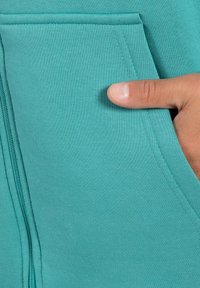 Felpa con zip color teal, dotata di tasca a marsupio. Il tessuto è liscio e presenta dettagli di cucitura visibili lungo la cucitura. Mano appoggiata sulla tasca.