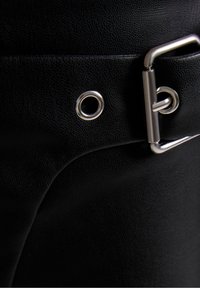 Svart läderaccessoar med en texturerad yta, som har en silverfärgad metallknäppning och en cirkulär ögla vid kanten.