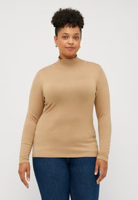 Beige langermet topp med høy hals og jevn tekstur, kombinert med blå jeans. Stoffet ser mykt og tettsittende ut, og fremhever silhuetten.