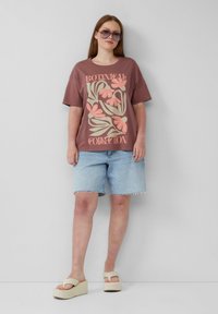 T-shirt graphique marron avec un design floral, associé à un short en denim bleu clair. Sandales à plateforme beiges avec détails de lanières. Lunettes de soleil brunes.