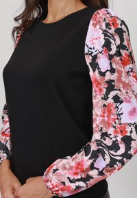 Sort topp med lange, mønstrede, puffede ermer med rosa og røde blomsterdesign på svart bakgrunn. Glatt materiale med tett passform.
