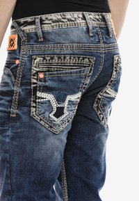 Denim-Jeans in einer verblassten blauen Farbe, mit kontrastierenden Nähten, dekorativen Gesäßtaschen und einem orangefarbenen Markenschild am Bund.