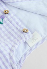 Tessuto a strisce lavanda e bianche con dettagli floreali ricamati, bottoni in legno e un'etichetta che recita "NEXT Baby." Texture morbida e design del colletto.