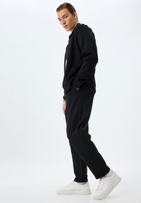 Sudadera negra con cremallera y cuello alto, combinada con pantalones jogger negros y zapatillas blancas. Tejido suave con un corte relajado.