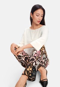 Weißes Langarmoberteil kombiniert mit gemusterten Hosen, die farbenfrohe Schmetterlingsmuster aufweisen; detaillierte Säume mit schwarzen Akzenten. Schwarze Schuhe runden das Outfit ab.