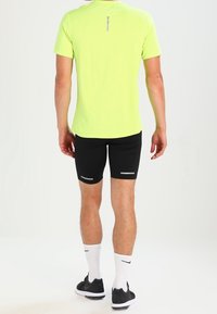 Camisa corta de manga amarilla brillante con detalles reflectantes, combinada con pantalones cortos negros ajustados que tienen acentos reflectantes y zapatillas deportivas negras.