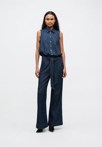 Denim jumpsuit with a sleeveless design, button-up front, belted waist, and wide-leg trousers. Dark blue color with a smooth texture.
