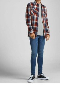 Camisa de franela a cuadros en tonos rojo, blanco y azul, con dos bolsillos en el pecho, llevada sobre una camiseta gris, combinada con jeans ajustados azules y zapatillas negras.