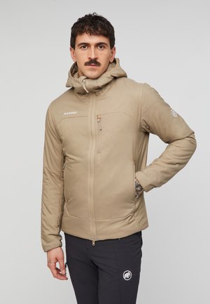 Mand med overskæg iført beige hættetrøje og sorte bukser, står med den ene hånd i lommen mod en ensfarvet baggrund.