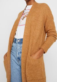 Modell som bär en karamellfärgad stickad cardigan över en vit grafisk t-shirt, ihop med högmidjade blå slitna jeans.
