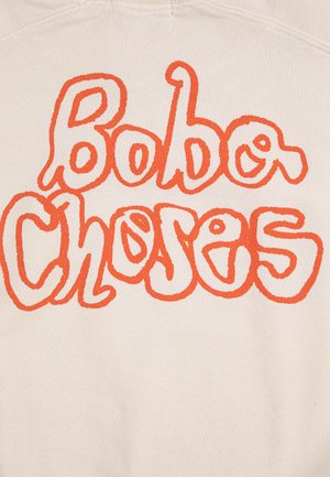 Beige stof med stor, bølget orange tekst, der læser "Bobo Choses" i legende, afrundet skrifttype.