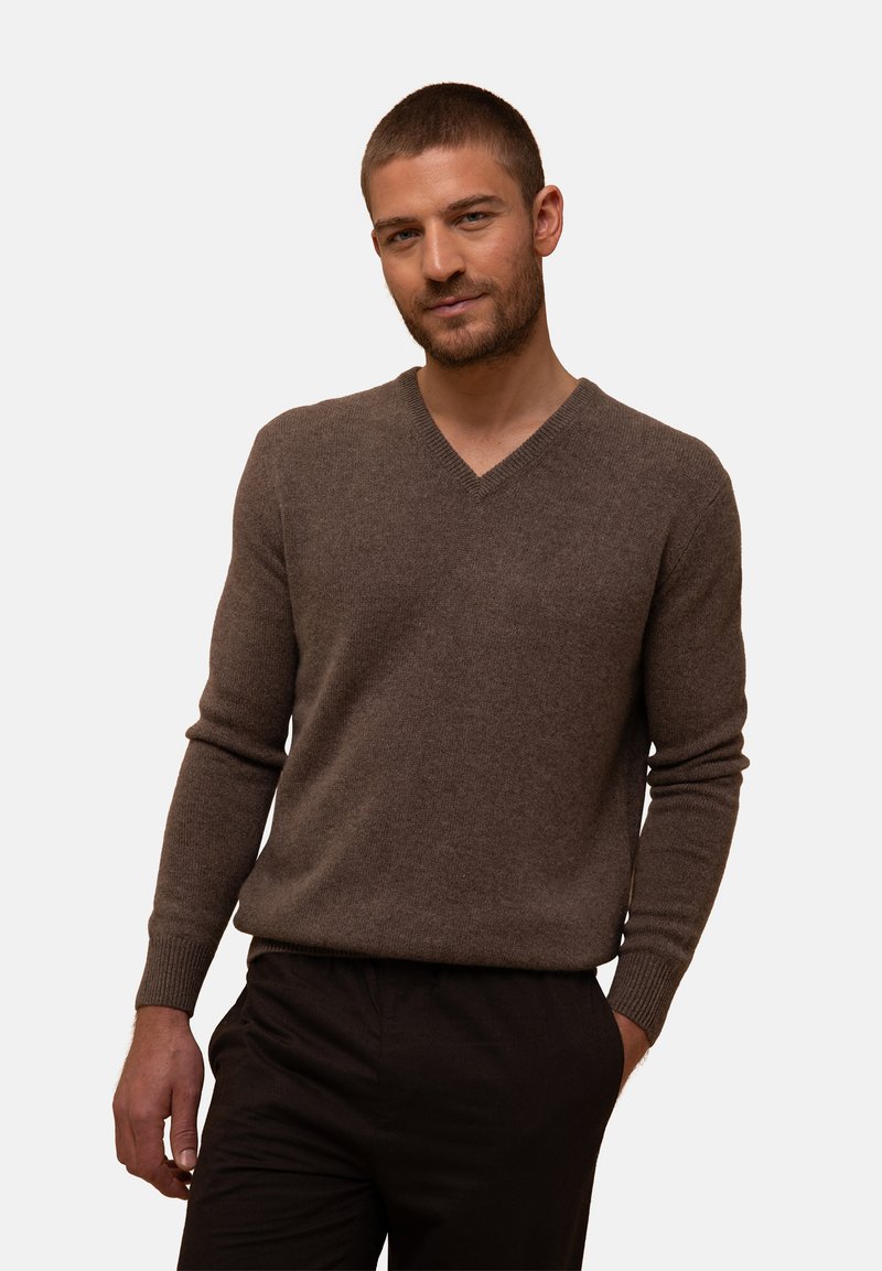 Brauner V-Ausschnitt-Pullover aus weicher, strukturierter Stoff. Lange Ärmel mit gerippten Bündchen. Kombiniert mit dunklen Hosen steht das Modell vor einem neutralen Hintergrund.