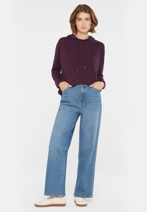 Frau steht mit Händen in den Taschen, trägt einen lila Kapuzenpullover, weite blaue Jeans und weiße Turnschuhe vor einem einfarbigen weißen Hintergrund.