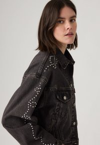 Young woman with dark hair wearing a black denim jacket adorned with silver studs in a zigzag pattern on the sleeve and shoulder.