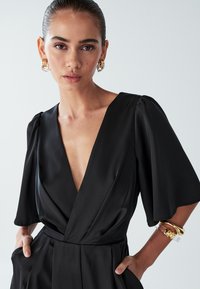 Sieviete ar nosmērētiem matiem, ģērbusies melnā kleitā ar dziļu V-veida izgriezumu, zeltainām auskaru gredzeniem un zeltainiem un caurspīdīgiem aproces, rokas kabatās, neitrālā fona.