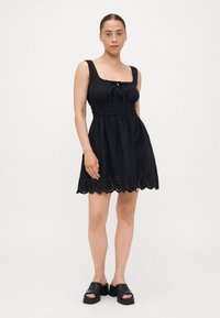 Μαύρο αμάνικο φόρεμα με τετράγωνη λαιμόκοψη, συγκεντρωμένη μέση και διακοσμητική ελαφριά δαντέλα στο στρίφωμα. Το ύφασμα φαίνεται ελαφρύ και λεία.