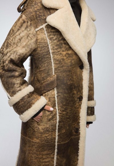 Brown shearling coat with beige lining, featuring a shawl collar, front pockets, and button closures. Textured finish with contrasting stitches.