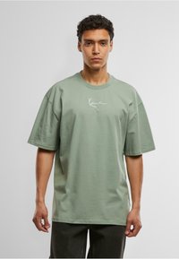 Green oversized t-shirt made from cotton, featuring a round neckline and short sleeves. Notable detail: white cursive logo on the chest.