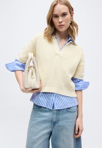 Γυναίκα που φοράει μπεζ πλεκτό πουλόβερ με κοντά μανίκια πάνω από μπλε ριγέ πουκάμισο, ανοιχτόχρωμο τζιν, κρατώντας μπεζ τσάντα με χρυστό φερμουάρ.