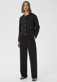 Svart kort jacka med guldknappar och frontfickor, kombinerad med vida svarta byxor, strukturerad tyg och enkel design.