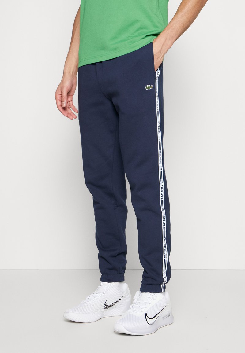 Pantalon de jogging navy avec une bande latérale blanche comportant du texte, fabriqué dans un tissu doux. Ceinture élastique, poches latérales, associé à des chaussures de sport blanches.