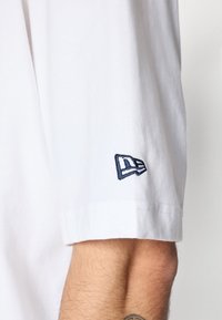 Camiseta de algodón blanca de manga corta. Presenta un logo bordado en navy en la manga. Textura suave y ajuste casual.